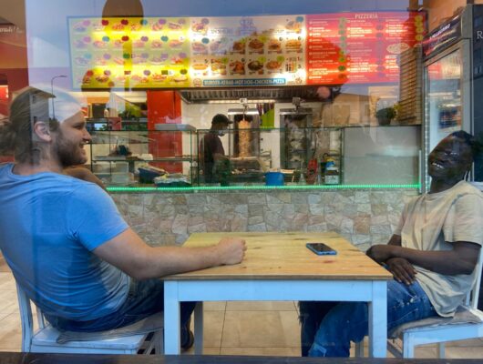 due ragazzi chiacchierano seduti a un tavolo, li vediamo attraverso un vetro di una finestra grande